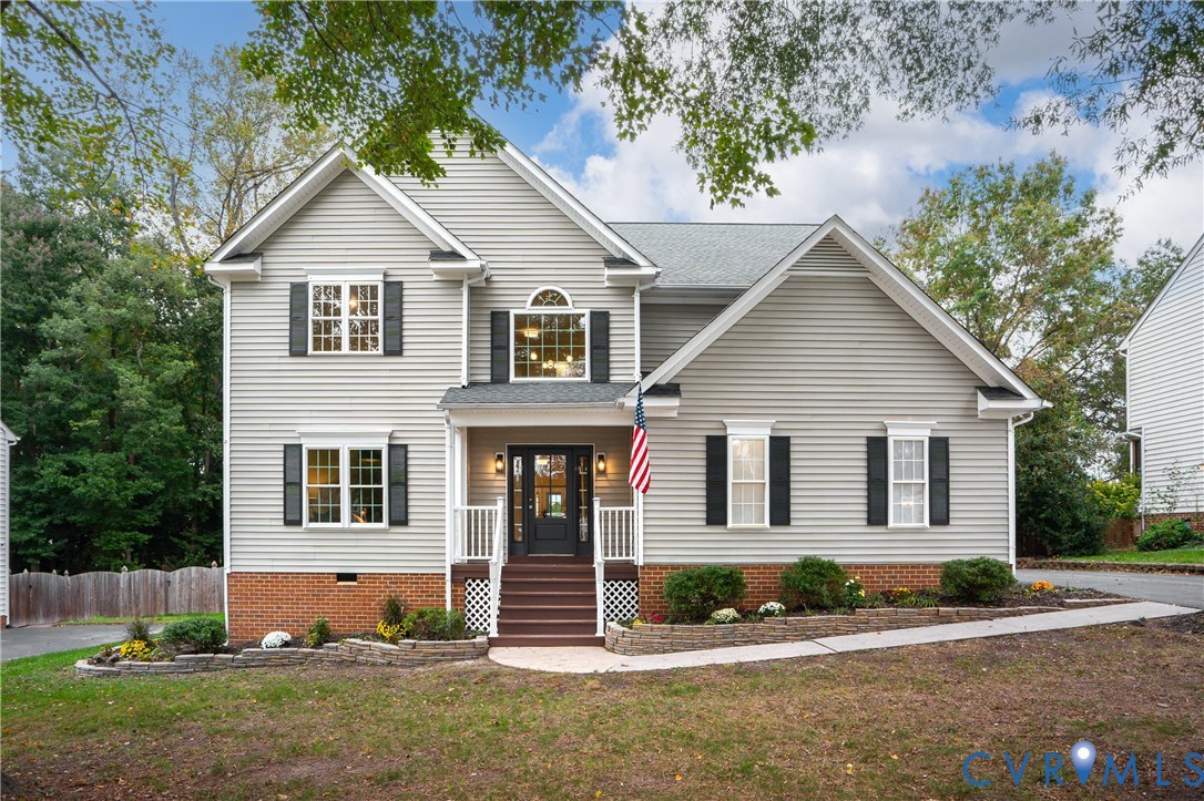 Two-story residence featuring light gray horizontal siding, brick foundation facade, multi-pane windows with black shutters, covered front porch with a wood-finished staircase, and tiered stone garden wall Two-story residence featuring light gray horizontal siding, brick foundation facade, multi-pane windows with black shutters, covered front porch with a wood-finished staircase, and tiered stone garden wall