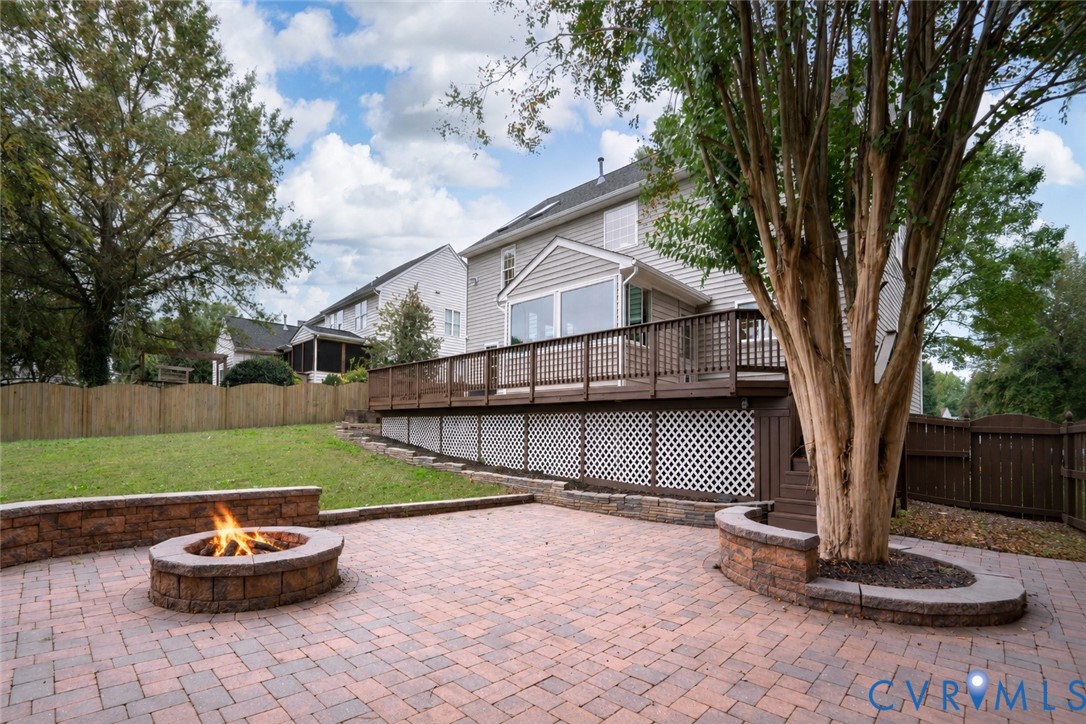 Backyard featuring a paved stone patio, built-in stone fire pit, tiered raised stone planter, elevated wood deck with lattice skirting, and wood privacy fencing