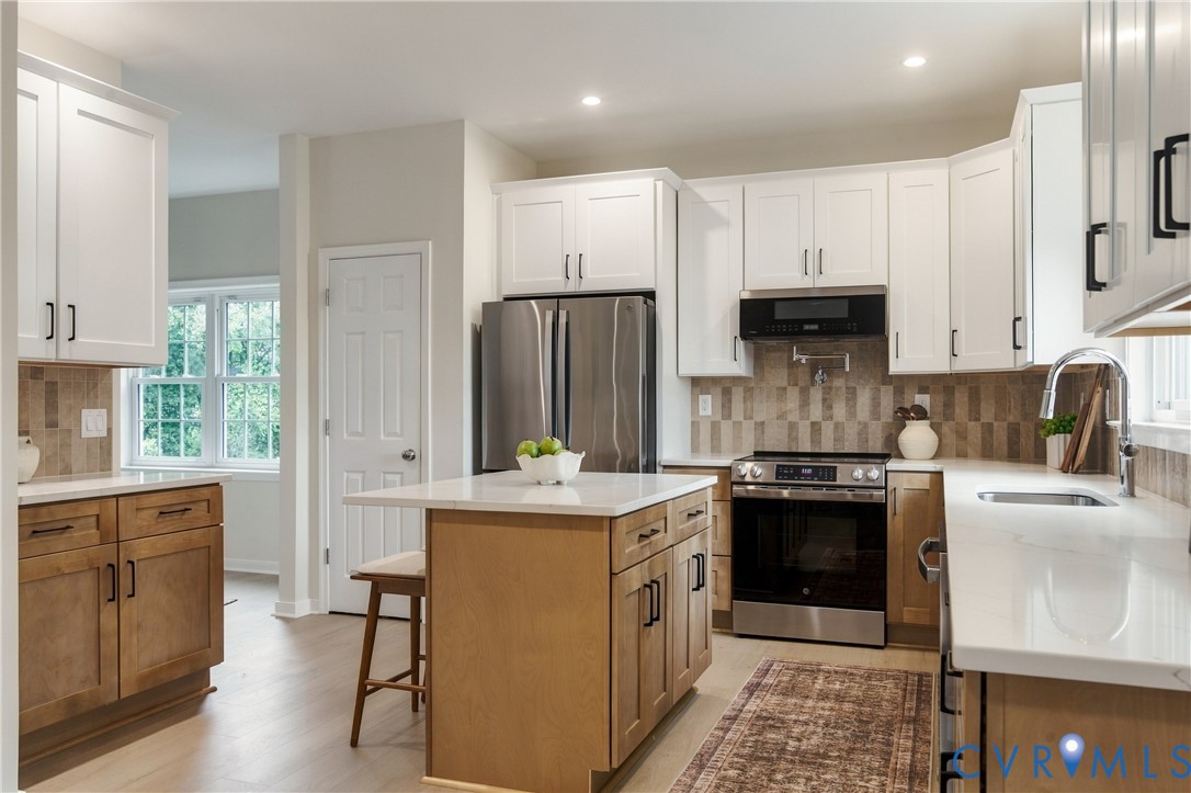 Kitchen featuring white shaker-style upper cabinetry, contrasting wood-finish base cabinets, a central island with a solid surface countertop, stainless steel appliances, and tile backsplash