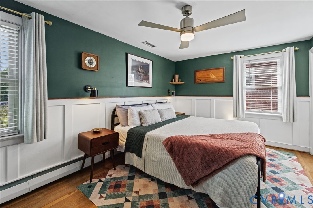 Bedroom featuring white wainscoting, matte dark green wall finish, wood-finish flooring, brushed nickel ceiling fan with integrated lighting, and dual windows