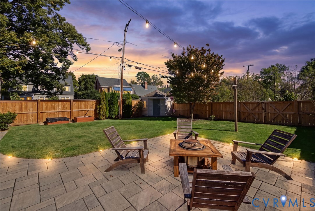 Fenced backyard featuring stone paver patio, manicured lawn, perimeter landscape lighting, and overhead string lights