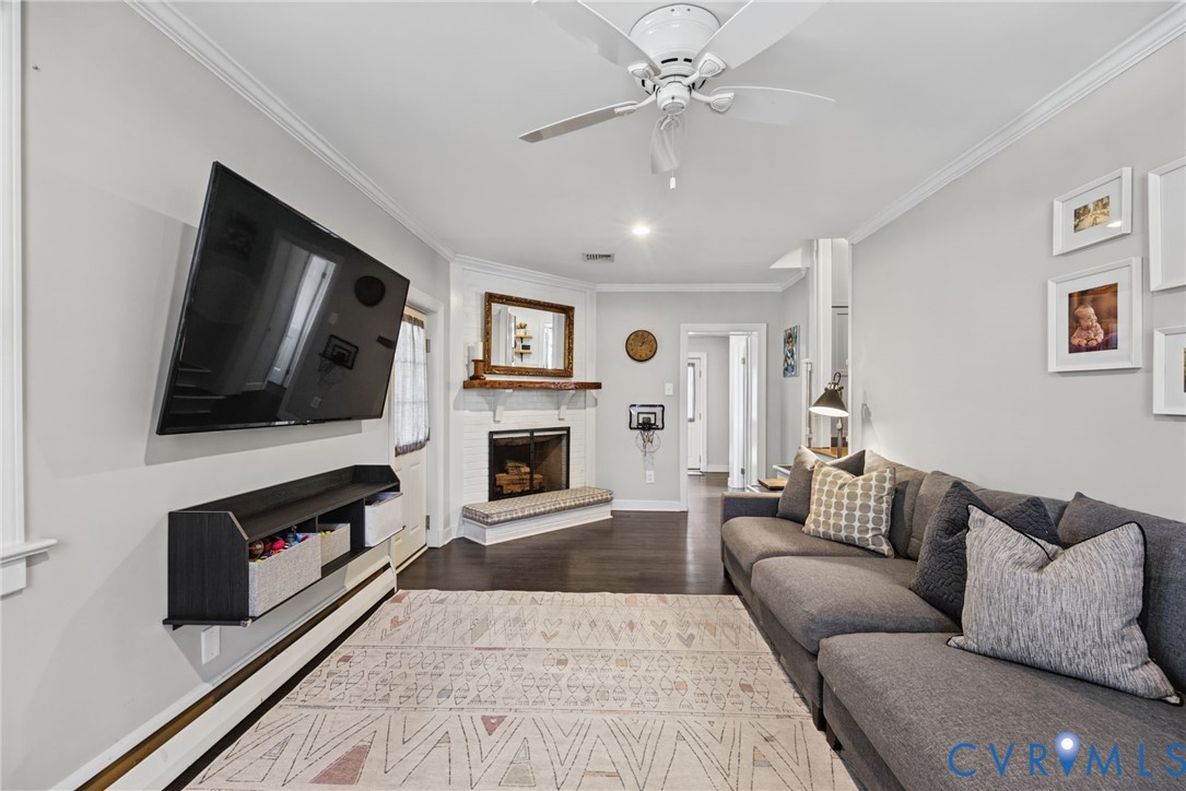 Corner fireplace featuring a painted brick hearth and wood mantel, white crown molding, wood-finish flooring, and a ceiling fan with integrated lighting