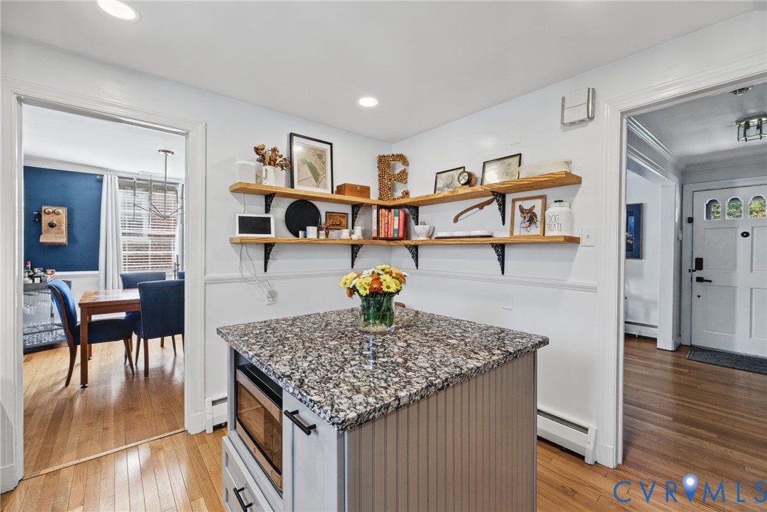 Kitchen island featuring stone-finish countertop, built-in microwave, and beadboard cabinetry, complemented by mounted natural wood shelving and wood-finish flooring