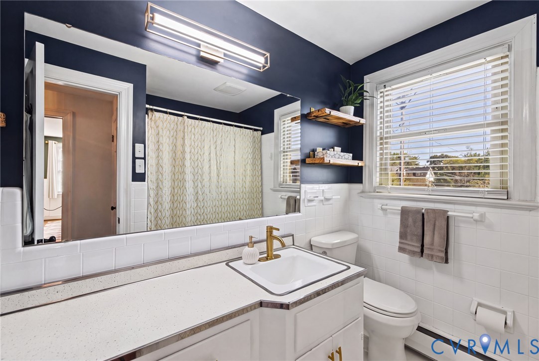 Bathroom featuring a white vanity with a solid-surface countertop, rectangular undermount sink with a brass-finish faucet, oversized wall-to-wall vanity mirror, and white tile wainscoting