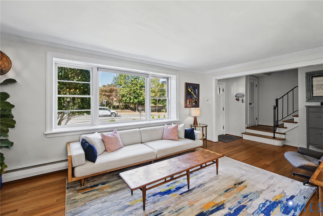 Living area featuring large multi-pane windows, wood-finish flooring, crown molding, and a staircase with a black metal railing Living area featuring large multi-pane windows, wood-finish flooring, crown molding, and a staircase with a black metal railing