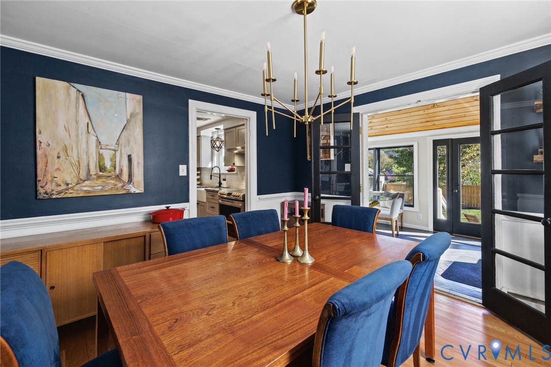 Dining room featuring dark blue walls with white wainscoting, wood-finish flooring, a multi-arm metallic chandelier, and glass-paneled french doors Dining room featuring dark blue walls with white wainscoting, wood-finish flooring, a multi-arm metallic chandelier, and glass-paneled french doors