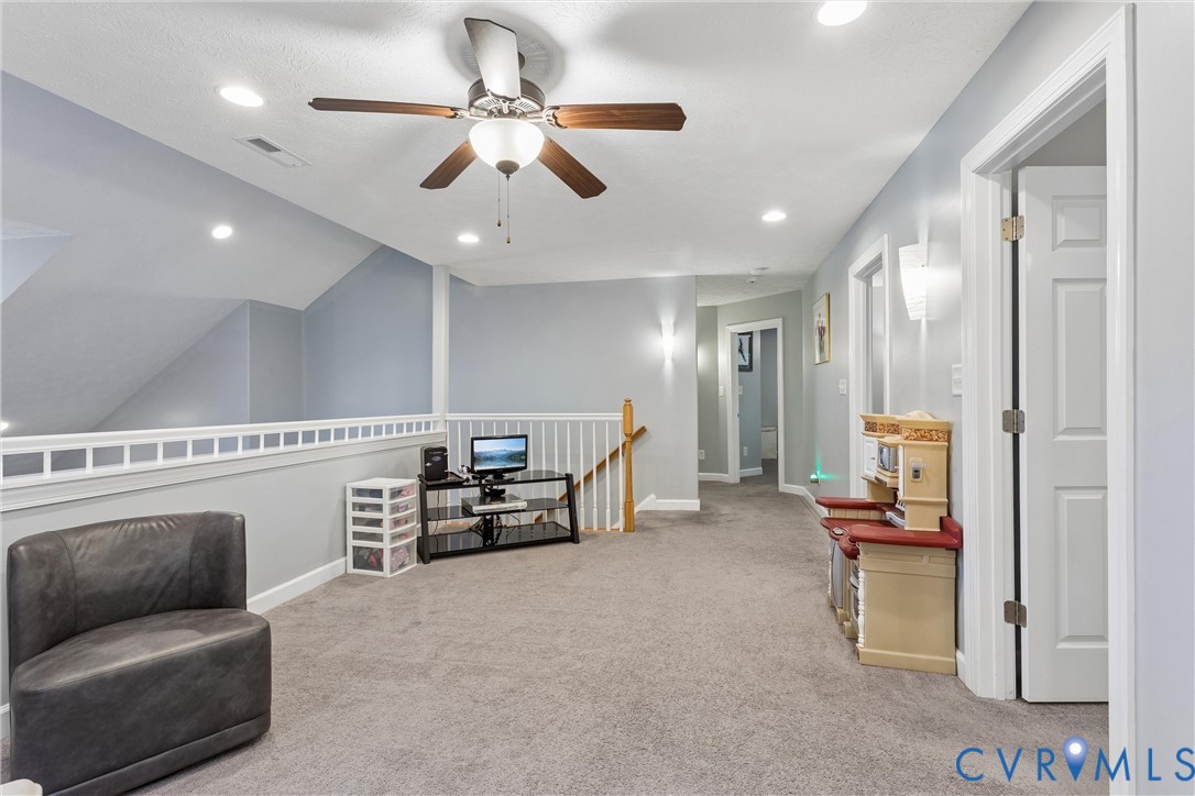 Neutral-toned carpeted interior featuring a ceiling fan with integrated lighting, recessed lighting, wall-mounted light fixtures, and a white balustrade railing