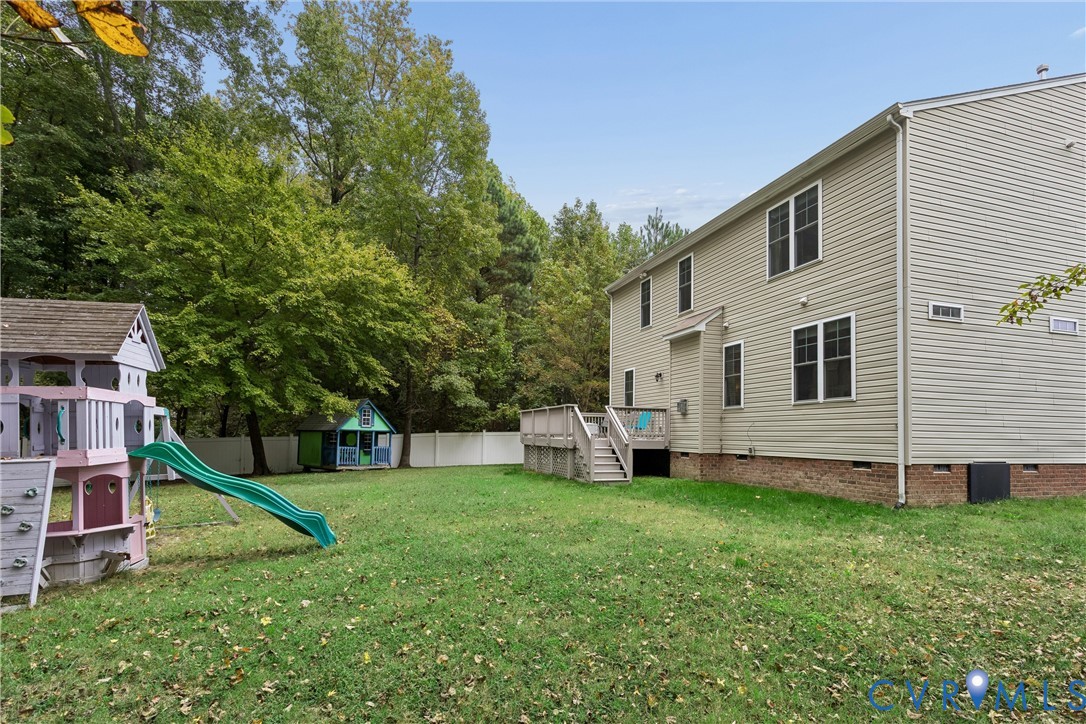 Expansive backyard featuring a leveled lawn, wood-toned elevated deck with stairs, full-perimeter white vinyl fencing, and mature perimeter trees