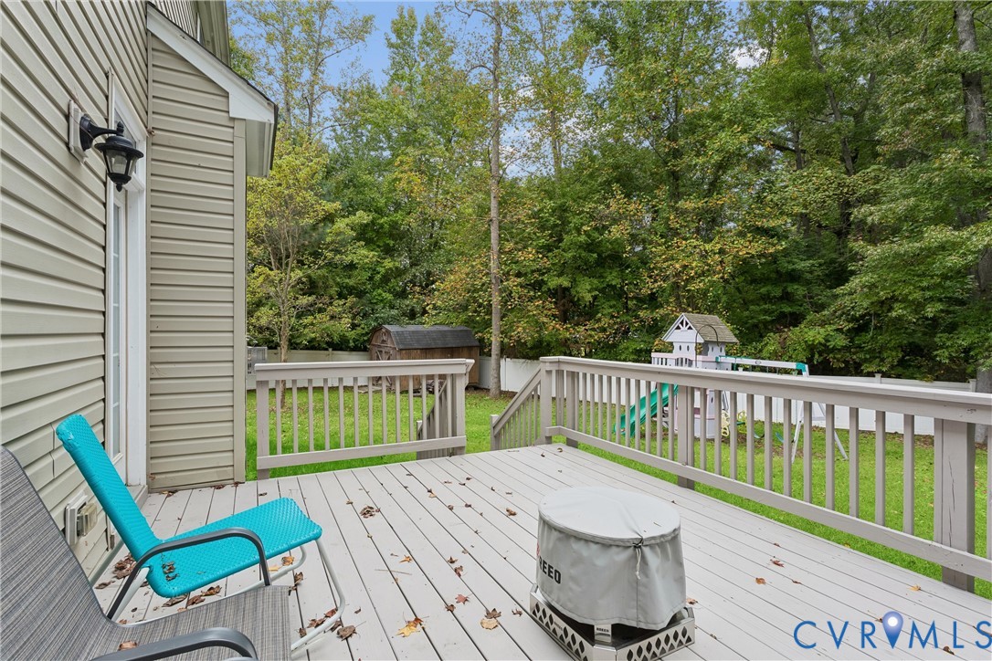 Raised wood-surface deck with perimeter railing, vinyl siding, wall-mounted exterior lighting, and fenced backyard with mature trees