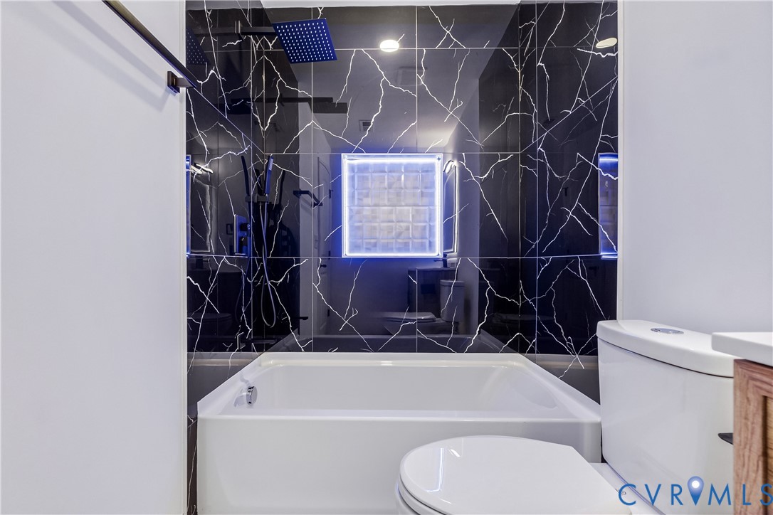 Bathtub surround featuring black patterned tile, rainfall showerhead, handheld shower fixture, and a glass block window with integrated illumination