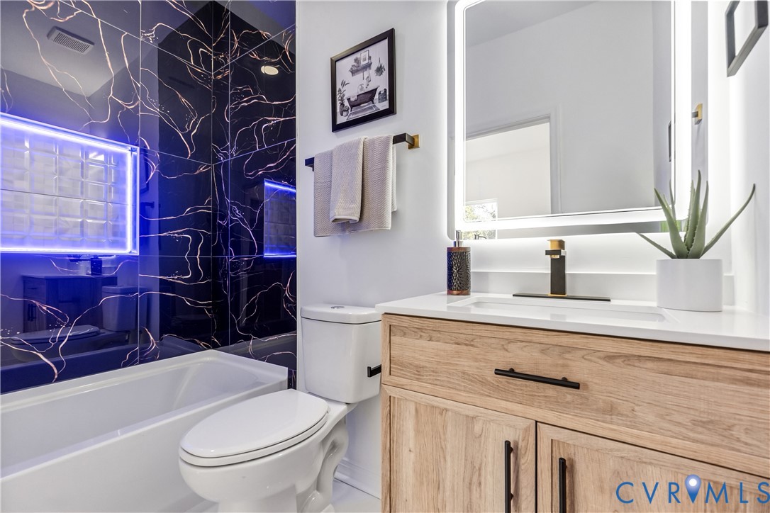 Bathroom featuring wood-finish cabinetry, a rectangular undermount sink with matte black hardware, a backlit wall-mounted mirror, and black wall tile with copper-toned veining in the shower area