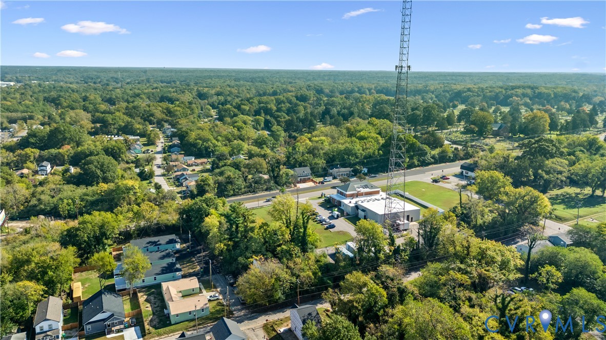 Aerial perspective featuring a commercial-style building with a flat roof and a prominent communications tower, situated on a lot with paved surface parking and surrounding mature tree canopy