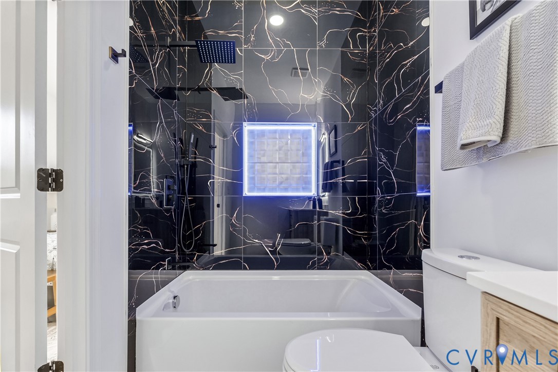 Bathroom featuring black vein-patterned wall tile, square rain-style shower head, integrated LED-lit glass block window, white soaking tub, and vanity with stone countertop