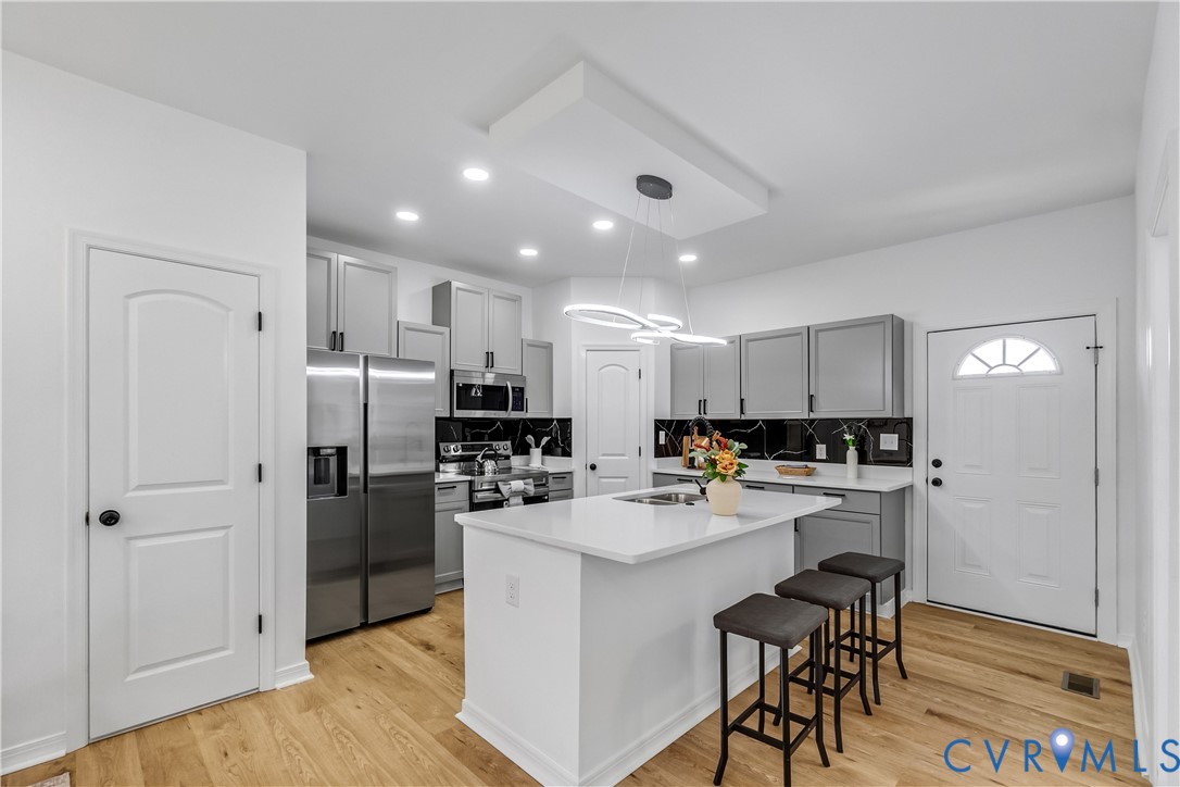 Kitchen featuring gray cabinetry, a white center island with integrated sink, stone-finish countertops, black tile backsplash, and wood-finish flooring