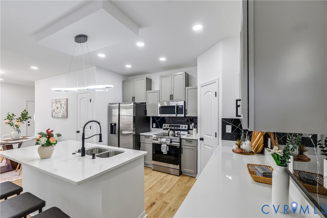 Kitchen featuring an expansive central island with a dual-basin undermount sink, quartz countertops, gray shaker-style cabinetry, stainless steel appliances, and wood-finish flooring