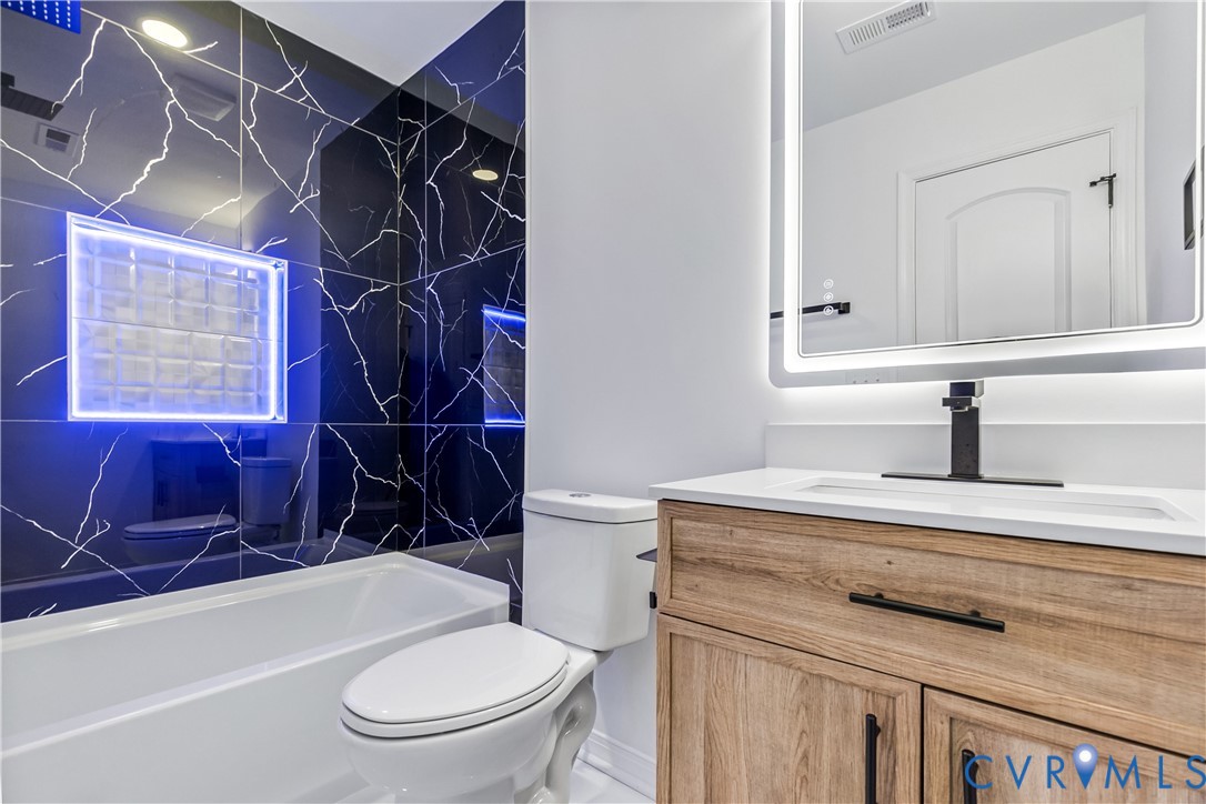 Bathroom featuring black tile with white veining, wood-finish vanity with white countertop, recessed glass block niche, and integrated LED-backlit mirror