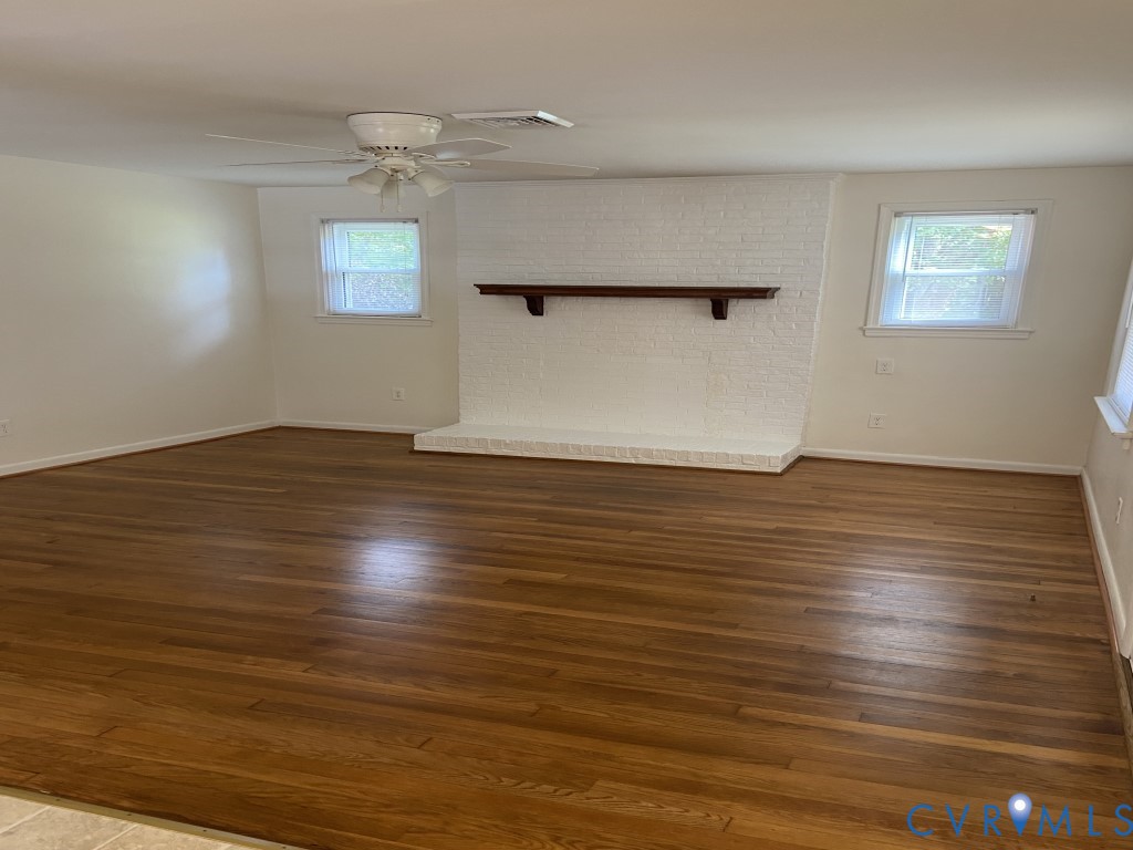 Painted brick fireplace with a wood mantel and hearth, wood-finish flooring, two windows with natural light, and a ceiling fan