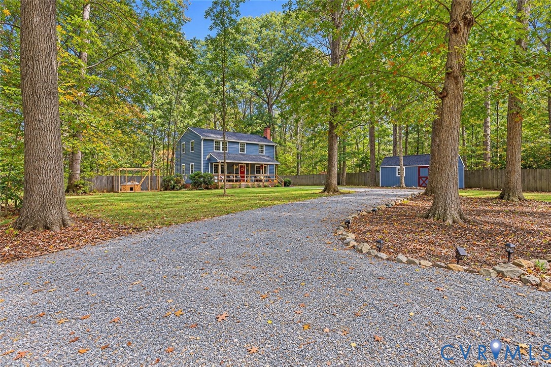 Gravel driveway leading to a two-story residence with a covered front porch, accompanied by a detached storage structure and expansive wooded grounds enclosed by rear fencing