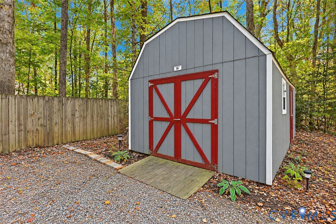 Detached storage shed featuring gray vertical siding, red double doors with cross-buck detailing, a wooden access ramp, and white trim