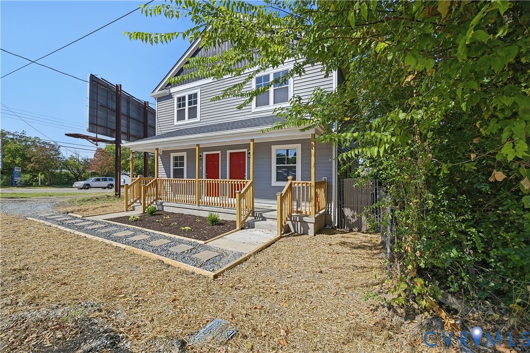 Two-story horizontal siding exterior with a covered front porch, wood-tone railings, and two individual red front doors, complemented by a front yard featuring a paved stone walkway and decorative mulch beds