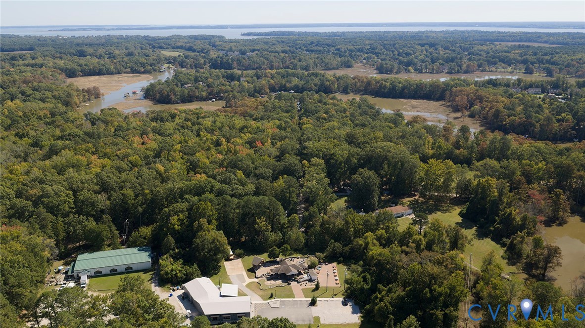 Aerial perspective of a sprawling property featuring multiple structures, paved hardscaping, a metal-roofed facility, and significant acreage with dense woodland and natural waterways