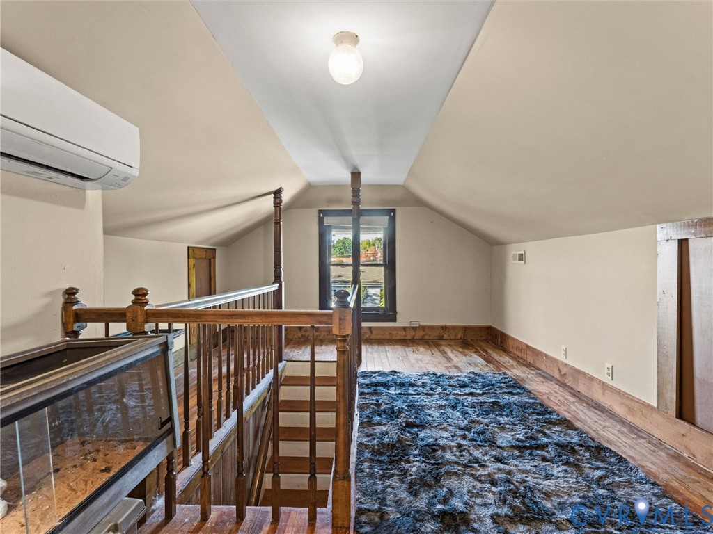 Vaulted ceilings, wood flooring, wood spindle railing, wall-mounted heating and cooling unit, and natural light from a window with dark wood trim