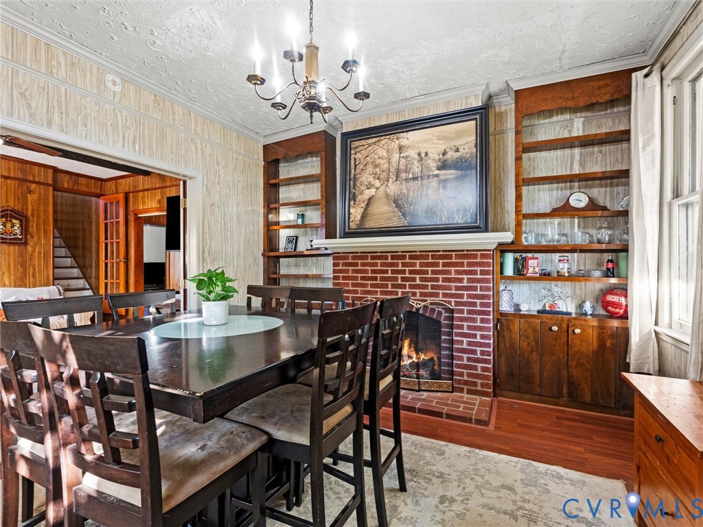 Dining area featuring a brick fireplace, built-in wooden shelving, a multi-arm chandelier, and wood-finish flooring. Flames are AI generated. Fireplace is not currently hooked to fuel source.