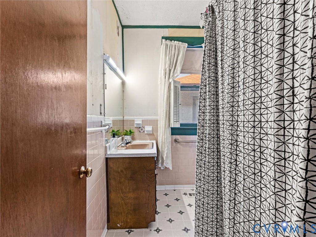 Bathroom featuring a wood-finish vanity with a rectangular sink, wall-mounted mirror, tiled wainscoting, and patterned vinyl flooring
