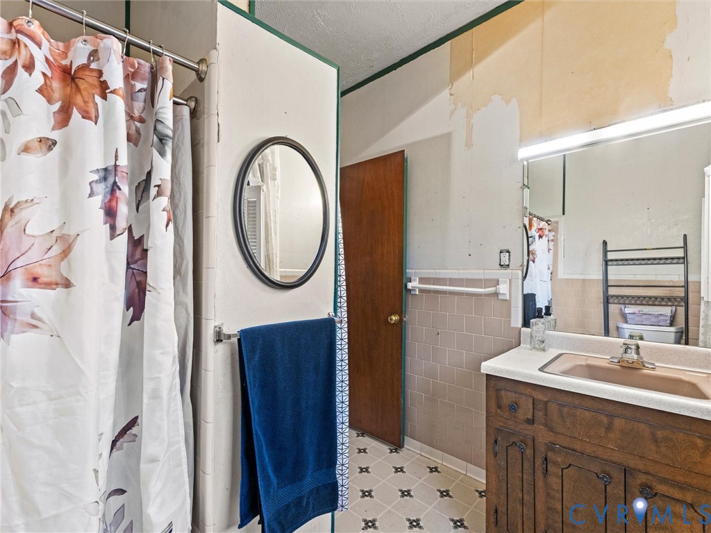 Bathroom featuring a wood-finish vanity with an integrated sink, large wall mirror, circular accent mirror, tiled wall accents, shower, and patterned vinyl flooring