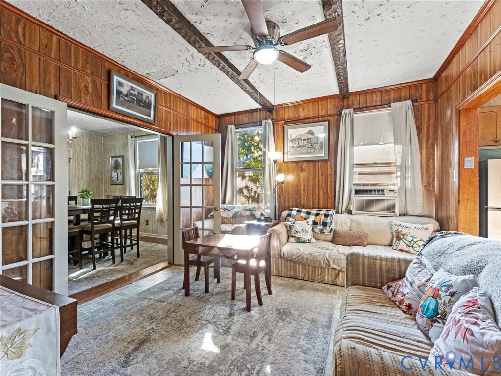 Living area featuring wood-paneled wall finish, exposed ceiling beams, multi-pane glass French doors, and ceiling fan with integrated lighting