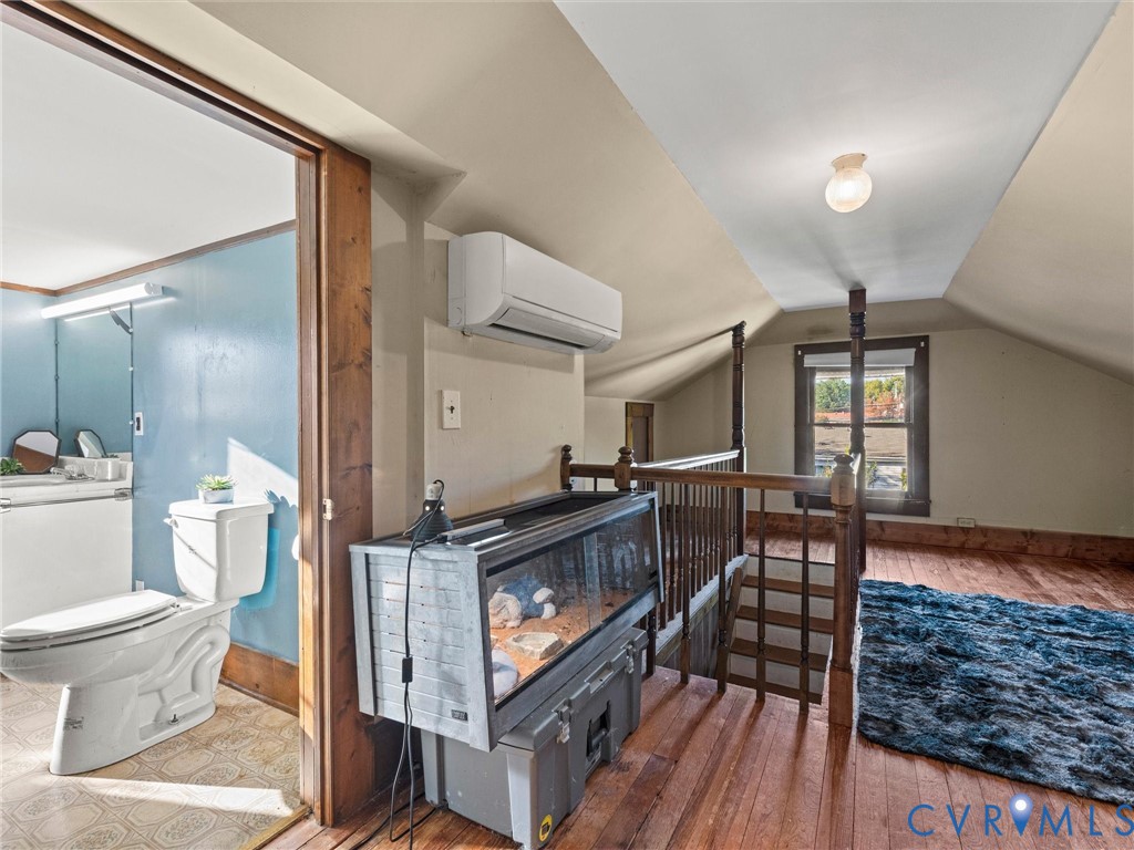 Sloped ceiling interior featuring wood flooring, a wall-mounted ductless HVAC unit, and a wooden balustrade staircase railing