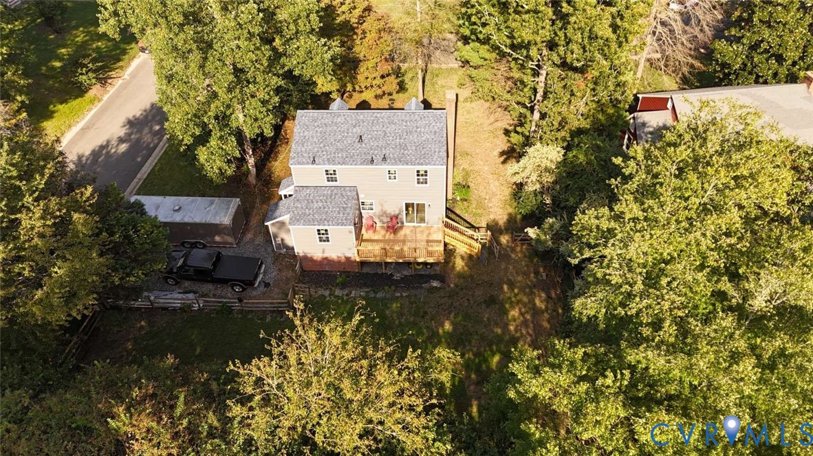 Aerial view of a two-story residence featuring an elevated rear deck, brick foundation, asphalt shingle roof, and a wooded lot