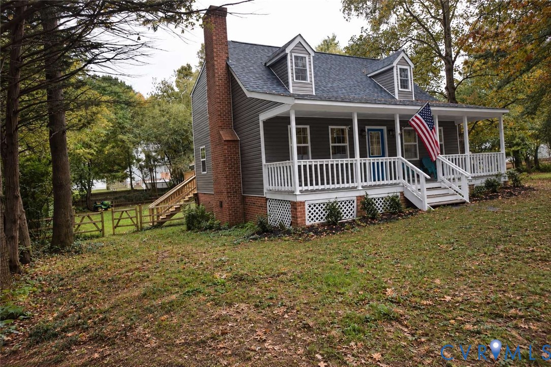 Cape Cod exterior featuring horizontal siding, a full-length covered front porch with white railing, a prominent exterior brick chimney, and dual dormer windows