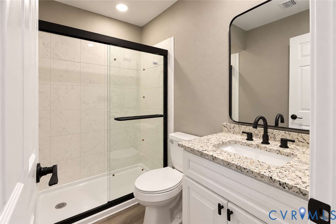 Bathroom featuring a walk-in shower with a glass enclosure and black metal frame, white cabinetry with speckled stone-finish countertop, undermount sink with matte black hardware, and a rounded rectangular wall mirror
