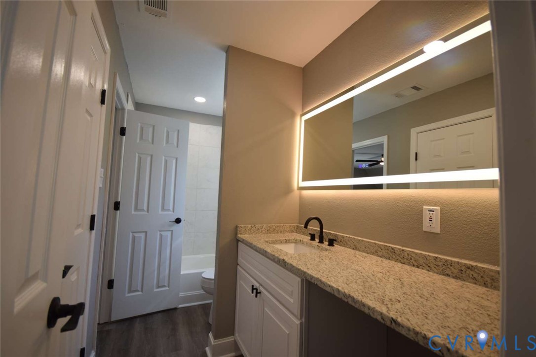 Bathroom featuring a vanity with stone-finish countertop, undermount sink, matte black faucet, and rectangular LED-backlit mirror, complemented by wood-finish flooring
