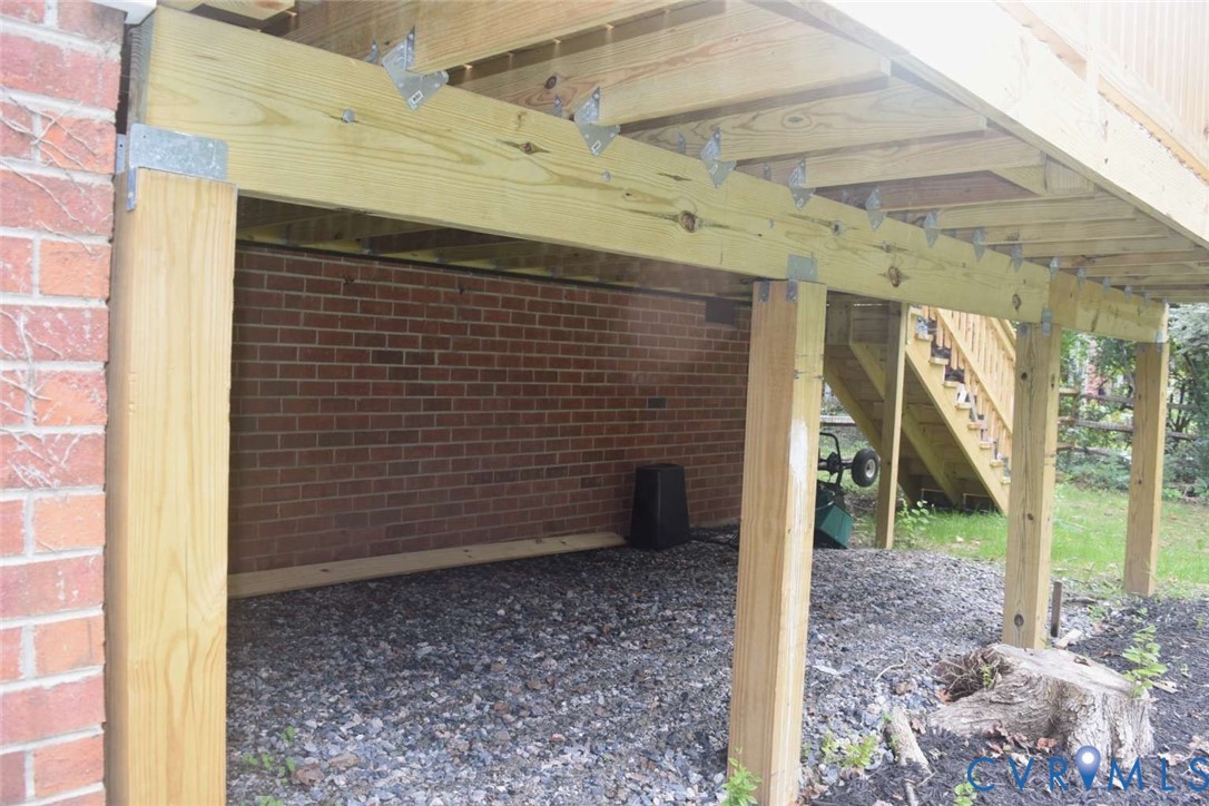 Raised wooden deck structure featuring structural support posts, joists, metal hardware connectors, and an attached staircase leading to a brick exterior wall with gravel ground covering