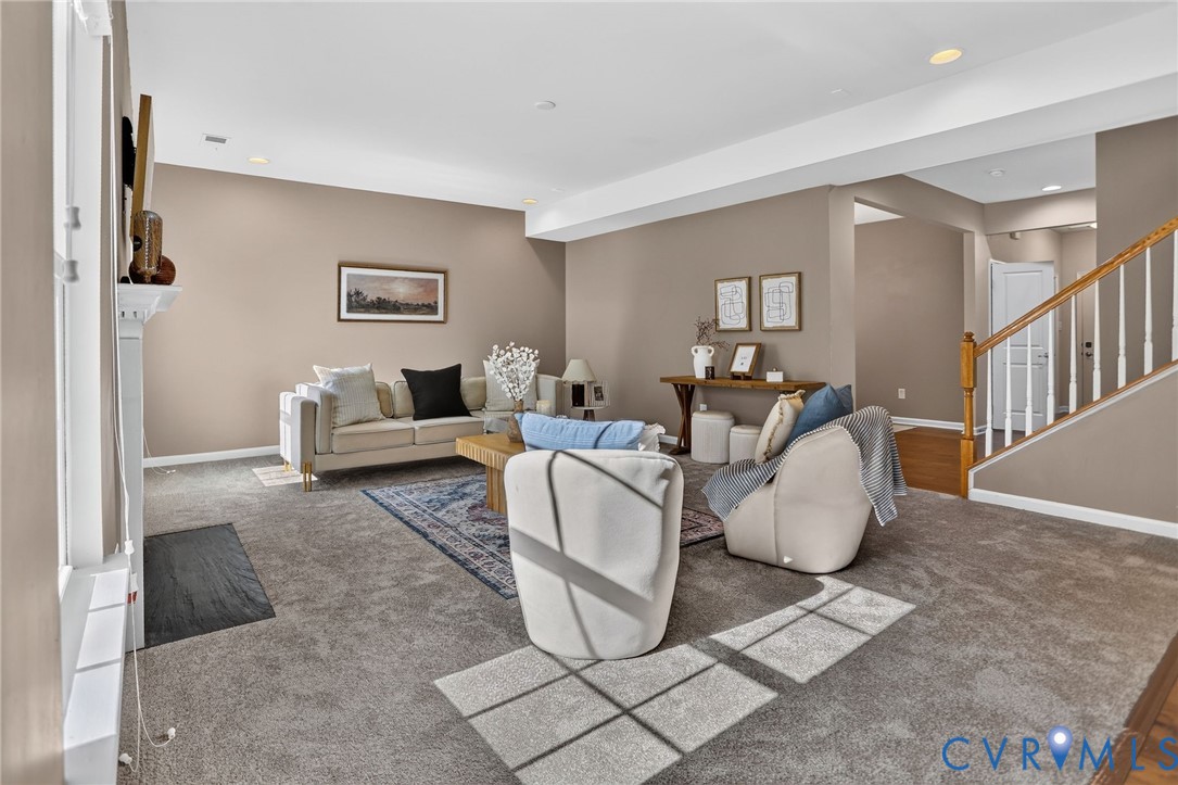 Open-concept interior featuring recessed lighting, a fireplace with a mantel, carpeted flooring, and a staircase with a wooden handrail Open-concept interior featuring recessed lighting, a fireplace with a mantel, carpeted flooring, and a staircase with a wooden handrail