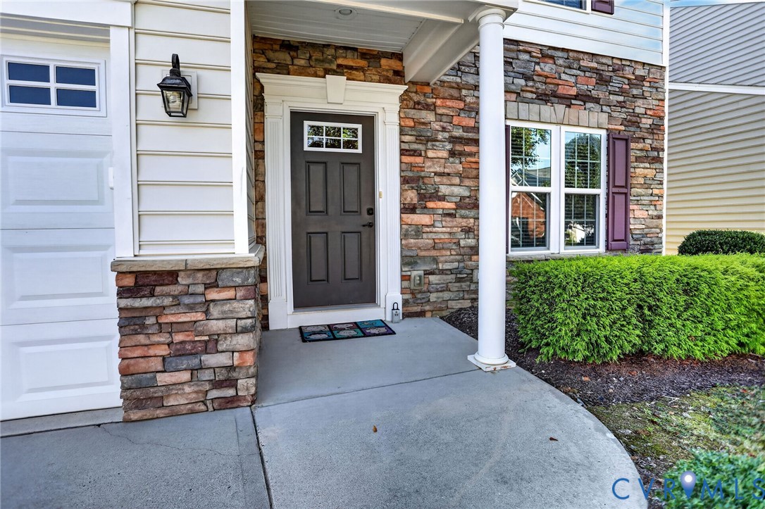 Exterior entry featuring a multi-tone stone veneer facade, horizontal vinyl siding, a covered concrete porch with a structural column, and a paneled front door with a transom window Exterior entry featuring a multi-tone stone veneer facade, horizontal vinyl siding, a covered concrete porch with a structural column, and a paneled front door with a transom window