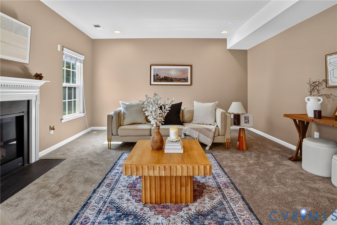 Neutral-toned living space featuring wall-to-wall carpeting, a fireplace with a decorative mantel, recessed ceiling lighting, and a window with trim detail Neutral-toned living space featuring wall-to-wall carpeting, a fireplace with a decorative mantel, recessed ceiling lighting, and a window with trim detail
