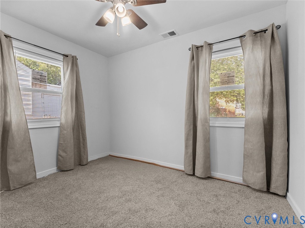 Room featuring neutral-toned carpeting, two windows with white trim, and a ceiling-mounted light fixture with a fan