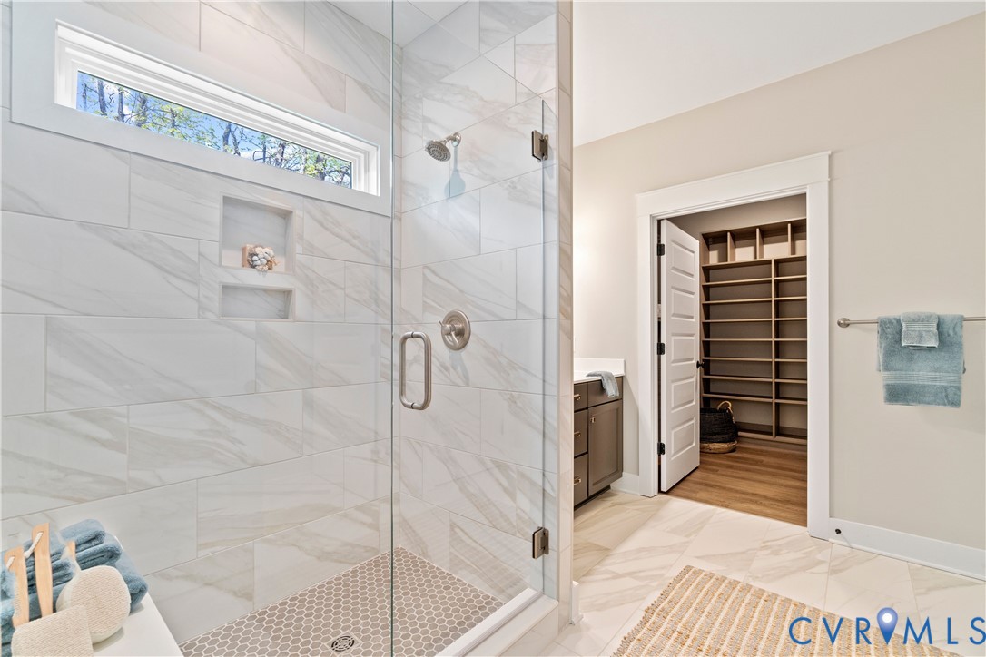 Walk-in shower featuring large-format tile surround, recessed wall niche, frameless glass enclosure, and circular mosaic tile flooring, accented by a walk-in storage area with custom built-in shelving