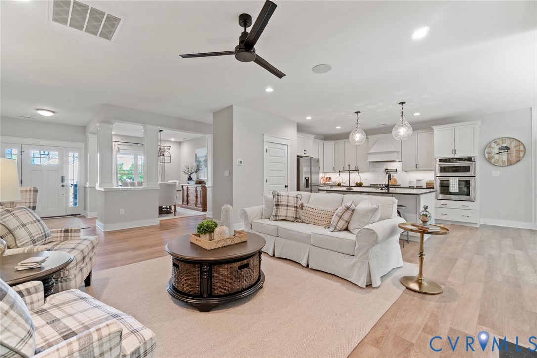 Open-concept interior featuring wood-finish flooring, a kitchen with white cabinetry and a central island, built-in double wall ovens, and a modern ceiling fan