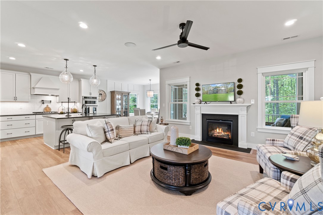 Open-concept living space featuring wood-finish flooring, recessed lighting, a ceiling fan, a kitchen with white cabinetry and an island, and a fireplace with a white mantel