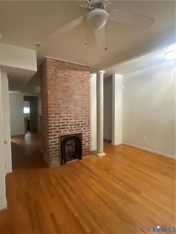 Exposed brick chimney breast with an arched metal fireplace surround, wood-finish flooring, decorative column, and ceiling fan with integrated lighting