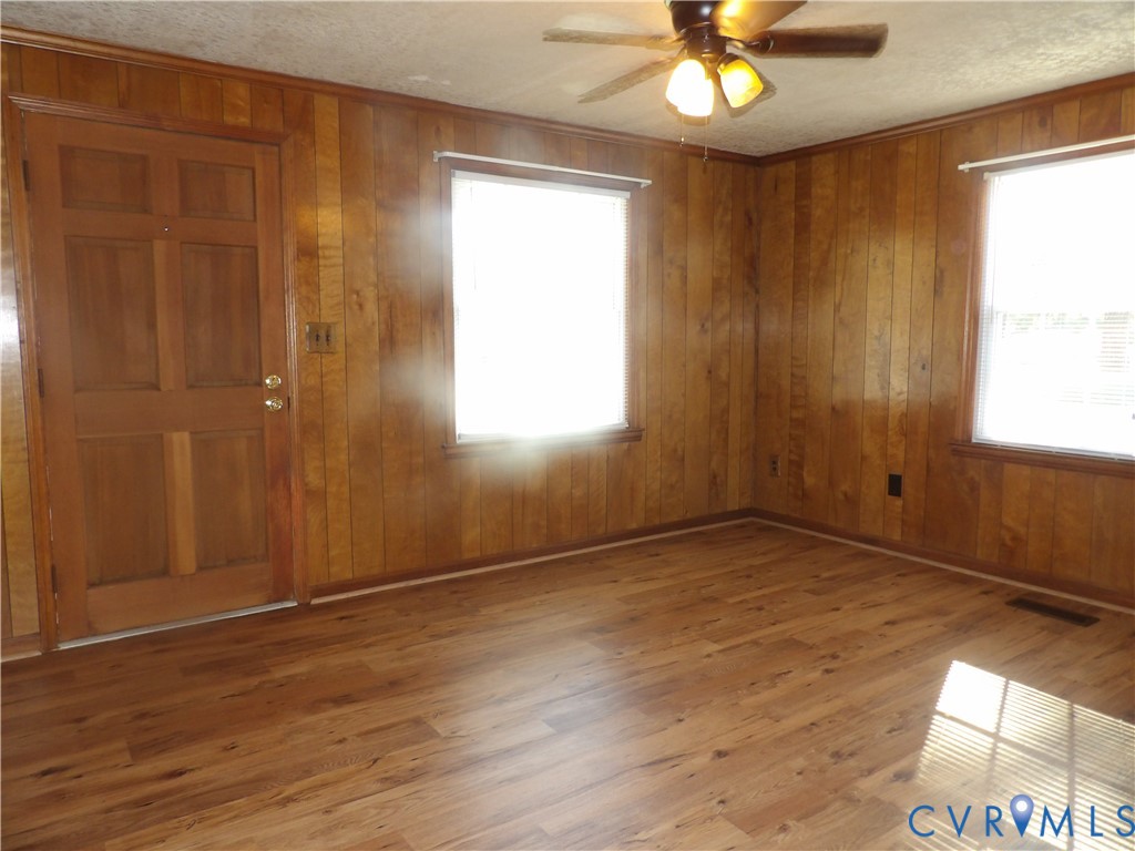Interior space featuring wood-panel wall treatments, wood-finish flooring, two large windows with horizontal blinds, a six-panel wood door, and a ceiling fan with light fixtures