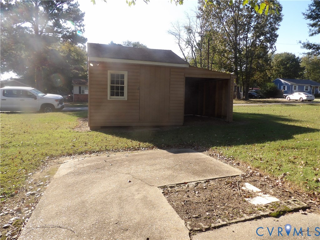 Detached outbuilding featuring horizontal wood-style siding, a gable roof, a single window, and an integrated open-air storage bay, complemented by a concrete pad