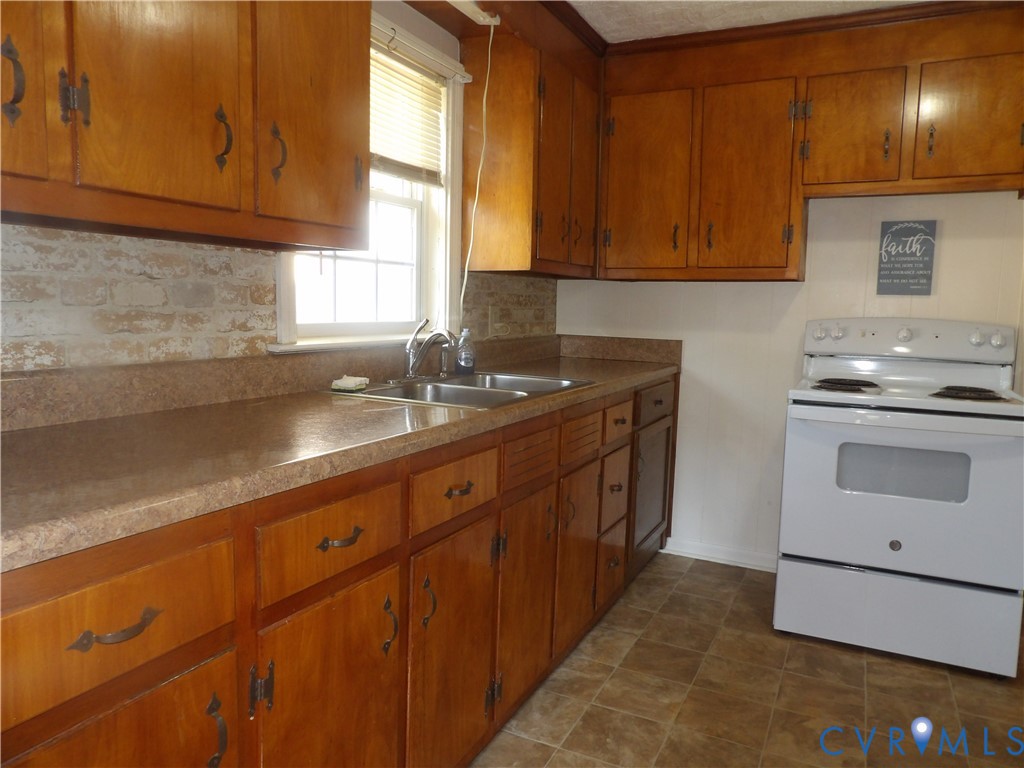 Kitchen featuring wood cabinetry, integrated countertops with a double-basin sink, exposed brick-style backsplash, and a white freestanding electric range