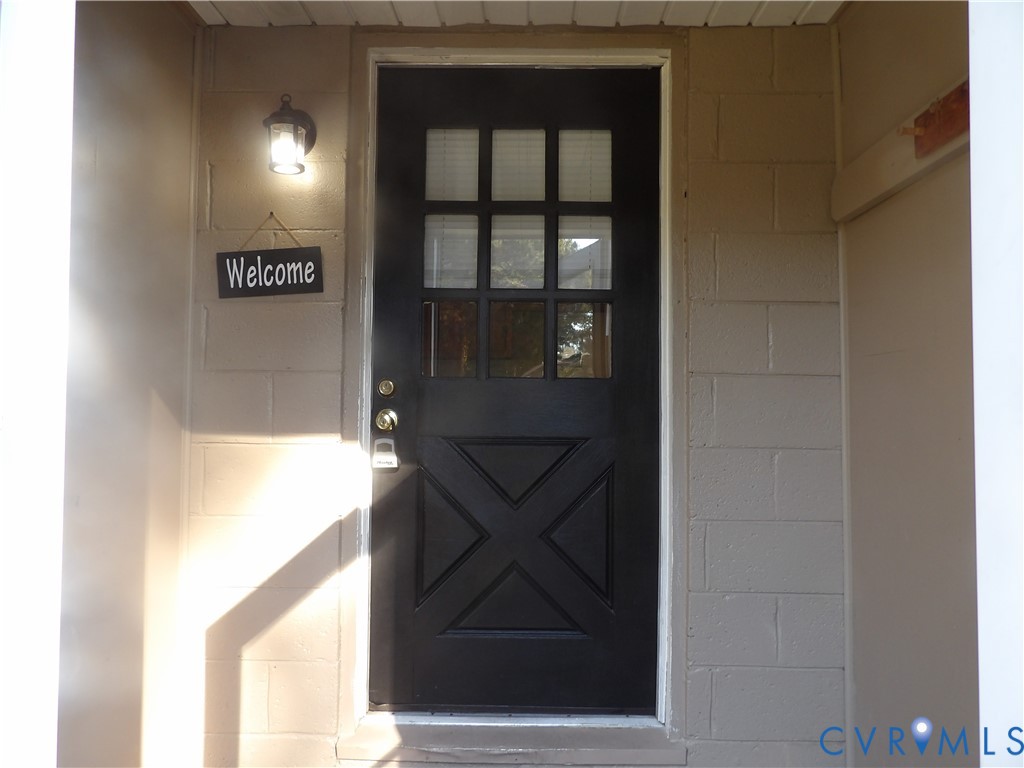Black exterior door featuring a nine-pane window inset and decorative lower cross-paneling, framed by concrete block masonry and an attached lantern-style wall sconce