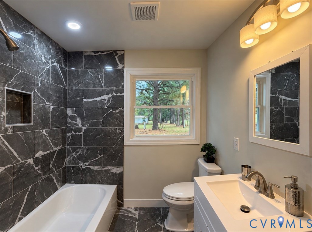 Bathroom featuring a deep soaking tub with large-format dark stone-finish tile surround, integrated shower niche, white vanity with rectangular basin, and wall-mounted vanity mirror with integrated lighting