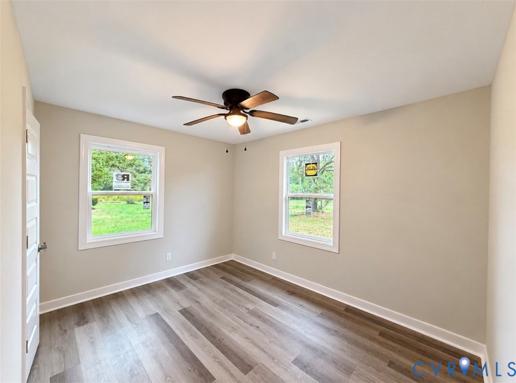 Neutral-toned room featuring wood-finish flooring, a ceiling fan with integrated lighting, and dual windows with white casing